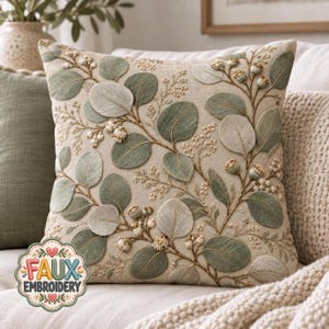 May include: A square throw pillow featuring an off-white linen-like fabric with detailed embroidery of sage green eucalyptus leaves, small white berries, and brown stems. A circular logo in the corner displays the text FAUX EMBROIDERY.