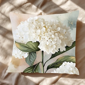 Könnte beinhalten: Quadratisches Zierkissen mit einer großen, detaillierten weißen Hortensienblüte mit grünen Blättern und einem hellbraunen Stiel. Der Hintergrund hat weiche Pastellfarben in Rosa, Beige und Blau. Das Kissen liegt auf einer beigen Oberfläche.