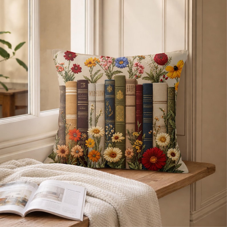 May include: A square throw pillow featuring a printed design of a row of vintage-style books standing upright. The books are interspersed with colorful illustrations of various wildflowers, including red, yellow, and white blossoms.