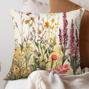 May include: Square decorative pillow featuring a watercolor floral design. The pillow showcases a field of wildflowers in shades of yellow, orange, pink, and purple against a cream background. The pillow is placed on a bed with other pillows and a blanket.