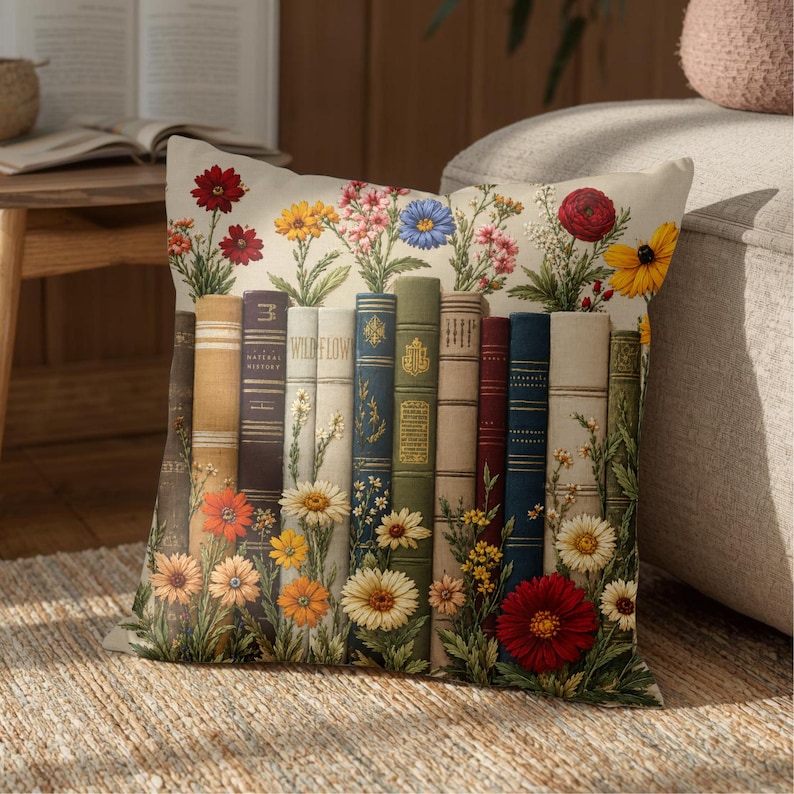 May include: A square throw pillow featuring a printed design of a row of vintage-style books standing upright. The books are interspersed with colorful illustrations of various wildflowers, including daisies, poppies, and sunflowers in shades of red, yellow, and blue.