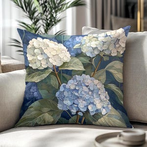 Può includere: Cuscino quadrato con un dipinto ad acquerello di fiori di ortensia blu e bianchi su uno sfondo blu scuro. Il cuscino ha foglie verdi e steli marroni. Il cuscino è su un divano grigio chiaro.