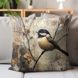 May include: Square cushion featuring a painted bird on a branch. The bird has a black head, brown and tan body, and a long black tail. The background is a textured, neutral colour with autumn leaves.