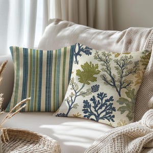 May include: Two decorative throw pillows on a cream-colored sofa. One pillow features vertical stripes in shades of blue and green. The other pillow has a botanical design with green and blue plant illustrations on a cream background.