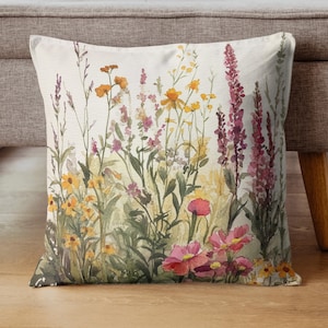 May include: Square throw pillow with a watercolor design of a wildflower meadow. The pillow features a variety of colorful flowers in shades of yellow, pink, and purple, set against a light background. The pillow is placed on a wooden floor.