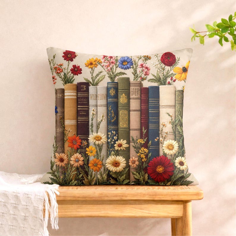 May include: A square throw pillow featuring a printed design of a row of vintage-style books standing upright. The books are interspersed with colorful illustrations of various wildflowers, including red, yellow, and blue blossoms, arranged at the base.