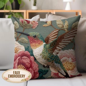 May include: Square throw pillow with a detailed embroidered design featuring a hummingbird with outstretched wings, surrounded by pink and red flowers and green leaves. The pillow has the words "FAUX EMBROIDERY" on it.