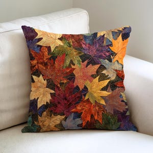 May include: Square throw pillow featuring a collage of colorful autumn leaves in shades of red, orange, yellow, and purple. The leaves are arranged closely together, creating a textured and vibrant pattern. The pillow is placed on a white sofa.