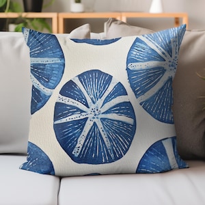 May include: Square throw pillow with a repeating pattern of blue and white circular designs. The blue designs have a watercolor effect, with radiating lines and a central starburst pattern. The background is a cream color.