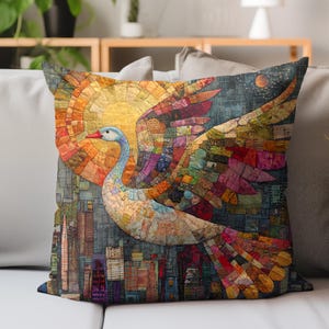 May include: Decorative throw pillow featuring a colorful mosaic design of a swan in flight. The swan has a blue neck and red beak, with wings spread against a backdrop of a sun and city skyline. The pillow is square and rests on a light gray sofa.
