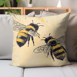 May include: A decorative throw pillow featuring two detailed illustrations of bees. The bees are black and yellow, with intricate wing and body details, set against a light yellow background. The pillow has a vintage, slightly distressed look.