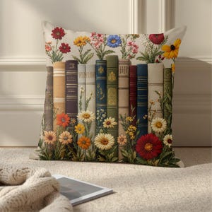 May include: A square throw pillow featuring a printed design of a row of upright books in various colors, including blue, green, brown, and cream. The books are interspersed with colorful illustrations of wildflowers, including red, yellow, and white daisies.