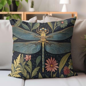 Könnte beinhalten: Dekoratives Zierkissen mit einer detaillierten Illustration einer Libelle mit blauen Flügeln und einem goldenen Körper. Das Kissen hat einen dunklen Hintergrund mit floralen und Blattakzenten in Grün-, Rosa- und Gelbtönen.