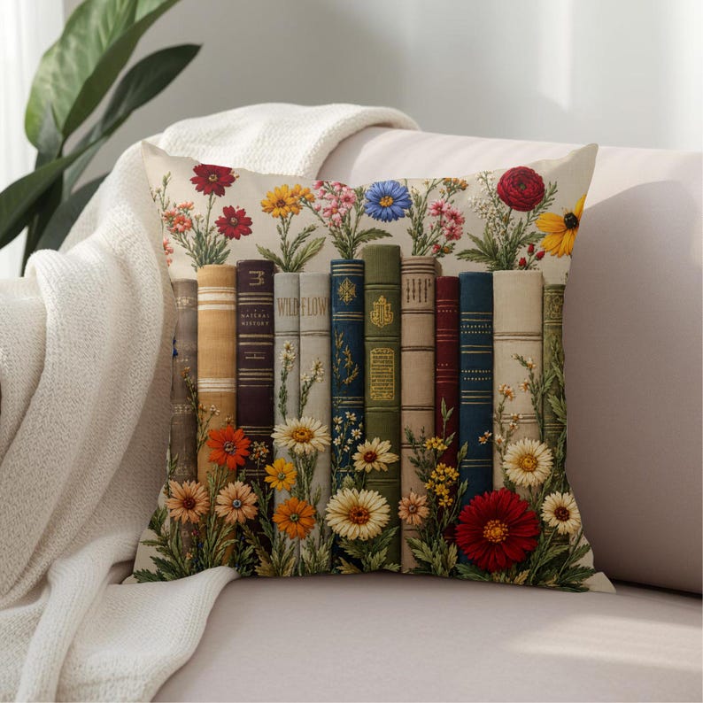 May include: A square throw pillow featuring a printed design of a row of vintage-style books in various colors including blue, green, brown, and cream, interspersed with colorful wildflowers like red, yellow, and orange daisies and small blossoms.