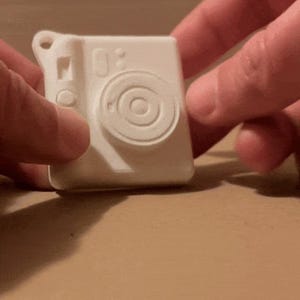 Könnte beinhalten: Eine weiße Spielzeugkamera aus Kunststoff mit einem Objektiv und einem Knopf an der Seite. Die Kamera ist etwa so groß wie eine Kreditkarte.