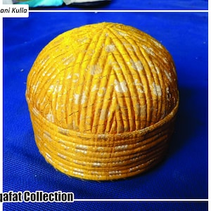 May include: A yellow Kulla hat with a patterned design, featuring the text "Afghani Kulla" and "Saqafat Collection". The hat has a rounded crown and a structured brim. The fabric has a gold and white design. The hat is a traditional style.