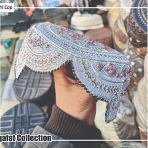 Peut inclure: Une casquette Sindhi argentée avec des broderies complexes et des ornements colorés. La casquette présente un motif détaillé avec des accents rouges, dorés et autres teintes de bijoux. Le texte "Sindhi Cap" et "Saqafat Collection" sont visibles.