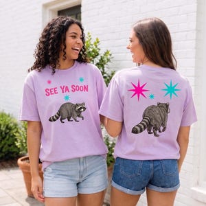 Puede incluir: Dos camisetas de color lila claro con un diseño de mapache. La parte delantera de la camiseta dice "SEE YA SOON" con una ilustración de mapache. La parte trasera de la camiseta tiene un gráfico de mapache y diseños de estrellas en rosa y turquesa.