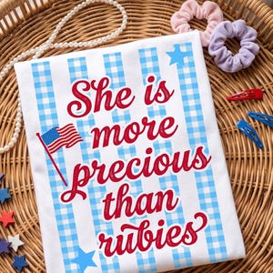 Puede incluir: Camiseta blanca con estampado de cuadros azules y texto "She is more precious than rubies" en rojo. La camiseta presenta una bandera americana y estrellas azules. Accesorios: coleteros, pinzas para el pelo y collar de perlas.