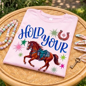 Puede incluir: Una camiseta rosa claro con las palabras "HOLD YOUR" en escritura azul. Se presenta un caballo marrón con una silla de montar turquesa, junto con una herradura roja y diseños de estrellas. La camiseta se muestra sobre una mesa de mimbre.