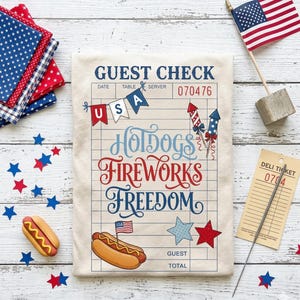 以下が含まれることがあります： 「Hotdogs, Fireworks, Freedom」の文字が入ったベージュ色のゲストチェックをメインとした愛国的な構成。チェックにはUSAバナー、ホットドッグのグラフィック、小さなアメリカ国旗が描かれています。星型の紙吹雪、おもちゃのホットドッグ、ミニチュアのアメリカ国旗が散りばめられています。