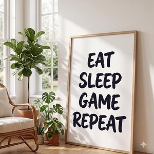 Op de afbeelding: Een ingelijste print met de tekst "EAT SLEEP GAME REPEAT" in zwarte penseelletters. De print heeft een lichte houten lijst en leunt tegen een muur. De kamer is licht, met planten en een rotan stoel, wat een ontspannen sfeer creëert.