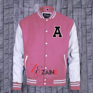 May include: Pink varsity jacket with white sleeves and a black letter "A" on the chest. The jacket has a button-up front, white stripes on the collar, cuffs, and hem, and the "Zaine Enterprises" logo.