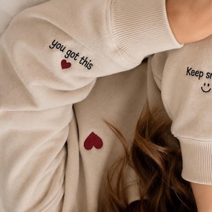 May include: Beige sweatshirt with the phrases "you got this" and "Keep smiling" embroidered in black. A red heart is embroidered near the phrase "you got this" and a smiling face is embroidered near the phrase "Keep smiling".