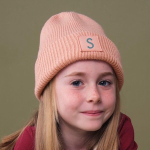 Op de afbeelding: Een perzikkleurige gebreide beanie met een vierkante patch met de blauwe letter "S". De beanie wordt gedragen door een persoon die een bordeauxrode longsleeve draagt. De achtergrond is een gedempt groen.