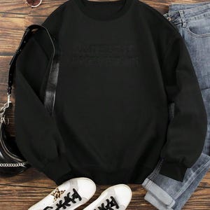 May include: Black sweatshirt with the text "ARTIST IN OPTIMIST BLACK". The long-sleeved top is paired with blue jeans, white sneakers with leopard print accents, a black purse, and sunglasses. The items are arranged on a wooden surface.