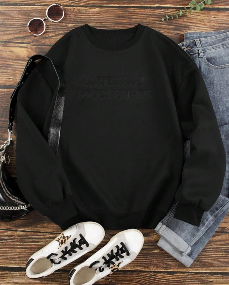 May include: Black sweatshirt with the text "ARTIST IN OPTIMIST BLACK". The long-sleeved top is paired with blue jeans, white sneakers with leopard print accents, a black purse, and sunglasses. The items are arranged on a wooden surface.