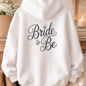 May include: A white hoodie featuring the phrase "Bride to Be" in black cursive script, with a diamond graphic. The sweatshirt is hanging on a wooden hanger. This item is likely intended for a bride-to-be, perfect for a hen party or wedding.