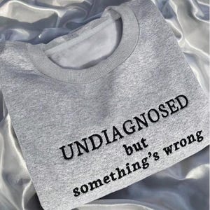 以下が含まれることがあります： 「UNDIAGNOSED but something's wrong」と黒い刺繍が施されたライトグレーのスウェットシャツ。クルーネックと長袖のデザインです。生地は柔らかく快適に見えます。