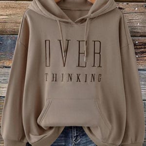 May include: A tan hoodie with the words "OVER THINKING" embroidered in dark brown. The hoodie has a front pocket, a hood with drawstrings, and long sleeves. The background is a wooden wall.