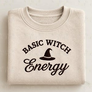 Könnte beinhalten: Ein beigefarbenes Sweatshirt mit Rundhalsausschnitt und dem schwarzen Schriftzug "BASIC WITCH", der über einem kleinen schwarzen Hexenhut-Symbol gebogen ist, sowie dem Wort "Energy" in Schreibschrift unter dem Hut. Das Kleidungsstück hat gerippte Bündchen.