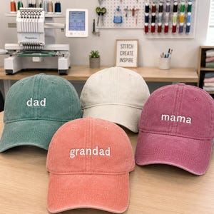 May include: Four baseball caps in green, beige, coral, and maroon. Each cap has a white embroidered word: "dad", "mama", and "grandad". The caps are displayed on a wooden surface.