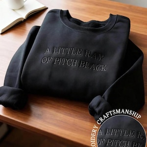 May include: Black sweatshirt with the embroidered text "A LITTLE RAY OF PITCH BLACK". The sweatshirt is folded on a wooden surface, with a white mug and an open book in the background. The image also includes a close-up of the embroidery.
