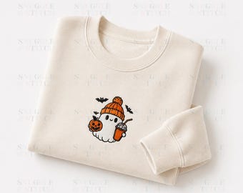 May include: A cream-coloured crewneck sweatshirt featuring an embroidered design of a white ghost wearing an orange beanie, holding a small orange pumpkin and a coffee cup, surrounded by three small black bats against a light background.