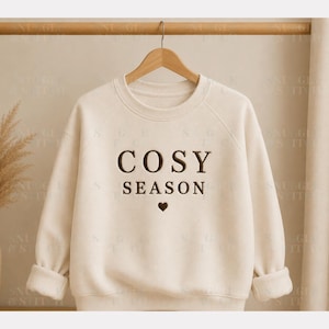 Pode incluir: Uma camisola de gola redonda cor de creme com o texto COSY SEASON bordado em castanho escuro no centro do peito, posicionado acima de um pequeno coração bordado em castanho escuro. A peça tem mangas compridas com punhos e gola canelados.