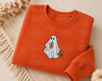 May include: A bright orange crewneck sweatshirt featuring an embroidered white ghost holding an iced coffee in one hand and a small orange jack-o'-lantern in the other. The ghost has black eyes and a mouth, with a green straw in the coffee cup.
