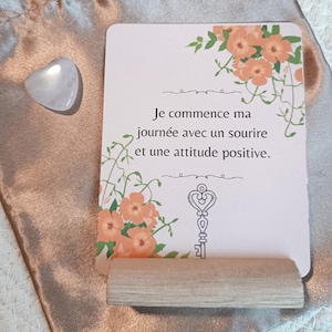 Peut inclure: Une carte rose clair avec le texte "Je commence ma journée avec un sourire et une attitude positive" et des motifs floraux. Un support en bois soutient la carte, une pierre en forme de cœur et une pochette en satin sont également visibles.