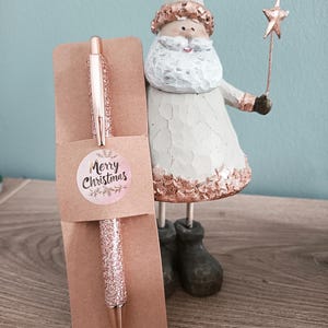 May include: A rose gold glitter pen in a brown paper sleeve with a "Merry Christmas" sticker. A decorative Santa Claus figurine with a star wand stands nearby. The pen is a festive gift idea.