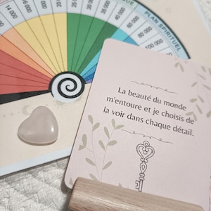 Peut inclure: Un cristal rose en forme de cœur à côté d'une carte avec du texte français et une illustration de clé. Un porte-cartes en bois et un graphique en forme de roue colorée sont également visibles.