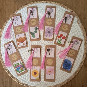 Peut inclure: Ensemble de marque-pages faits à la main avec des motifs floraux, chacun orné d'un pompon rose et d'un petit charme en forme de clé. Les marque-pages sont en papier kraft et portent l'inscription "handmade with love".