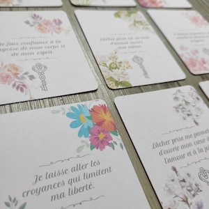 Peut inclure: Un jeu de cartes d'affirmation avec des motifs floraux et des citations inspirantes en français. Les cartes présentent un motif de trou de serrure et le texte "Je fais confiance à la sagesse de mon corps et de mon esprit." et "Je laisse aller les croyances qui limitent ma liberté."