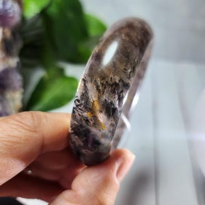 May include: A translucent, polished bracelet with a mix of dark brown, black, and yellow inclusions. The gemstone bracelet is held in a hand, showcasing its smooth, rounded form and natural patterns. The bracelet's design is organic and earthy.