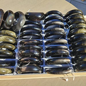 May include: A selection of dark, polished bangles in a wooden tray. The bracelets display a range of colours, including black, brown, and olive green, with a shiny surface. The bangles are arranged neatly, highlighting their smooth, curved shape. They appear to be made of stone or a similar material.