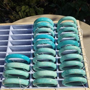 Op de afbeelding: Een display van jade armbanden in verschillende tinten turquoise en groen. De armbanden zijn gerangschikt in een grijze tray met individuele compartimenten. De belichting benadrukt de gladde, gepolijste oppervlakken van de sieraden en toont hun natuurlijke kleurvariaties.