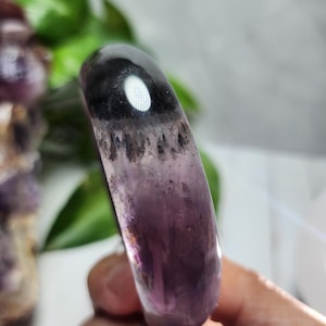 May include: A polished amethyst bracelet. The bracelet fades from a deep purple to black, with a white, reflective spot near the top. The stone has a smooth, rounded shape, and the light catches the internal patterns. The bracelet is approximately 2 cm wide.