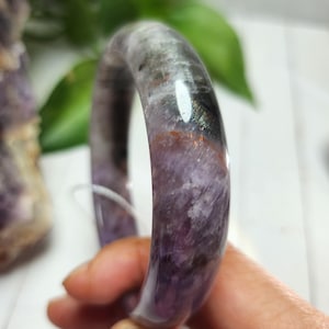 May include: A polished, translucent purple and grey gemstone bangle bracelet. The bracelet has a smooth, rounded shape and displays a mix of colours and patterns. The bracelet is held by a hand.